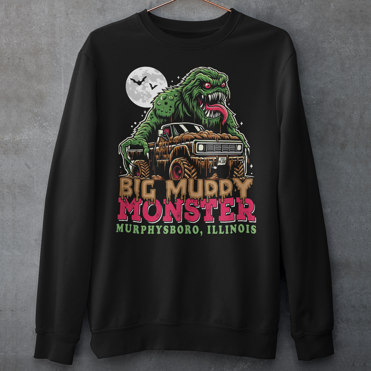 The sweatshirt design features the Murphysboro Mud Monster, a legendary cryptid, attacking a rugged, lifted truck. The monster, depicted with a hulking, muddy, and shaggy appearance, looms large over the vehicle, gripping the sides with massive, clawed hands. Mud splatters dramatically across the truck's tires and fenders, capturing the wild essence of the creature. Below the scene, the bold text "BIG MUDDY MONSTER" is prominently displayed.