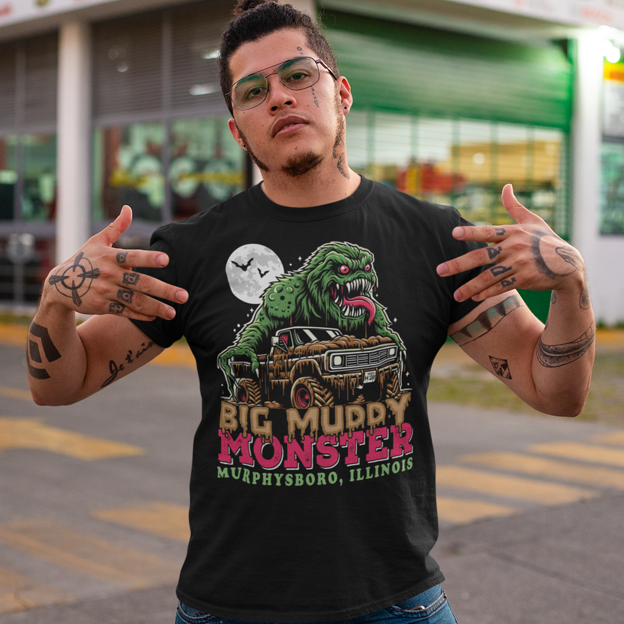 The t-shirt design features the Murphysboro Mud Monster, a legendary cryptid, attacking a rugged, lifted truck. The monster, depicted with a hulking, muddy, and shaggy appearance, looms large over the vehicle, gripping the sides with massive, clawed hands. Mud splatters dramatically across the truck's tires and fenders, capturing the wild essence of the creature. Below the scene, the bold text "BIG MUDDY MONSTER" is prominently displayed.