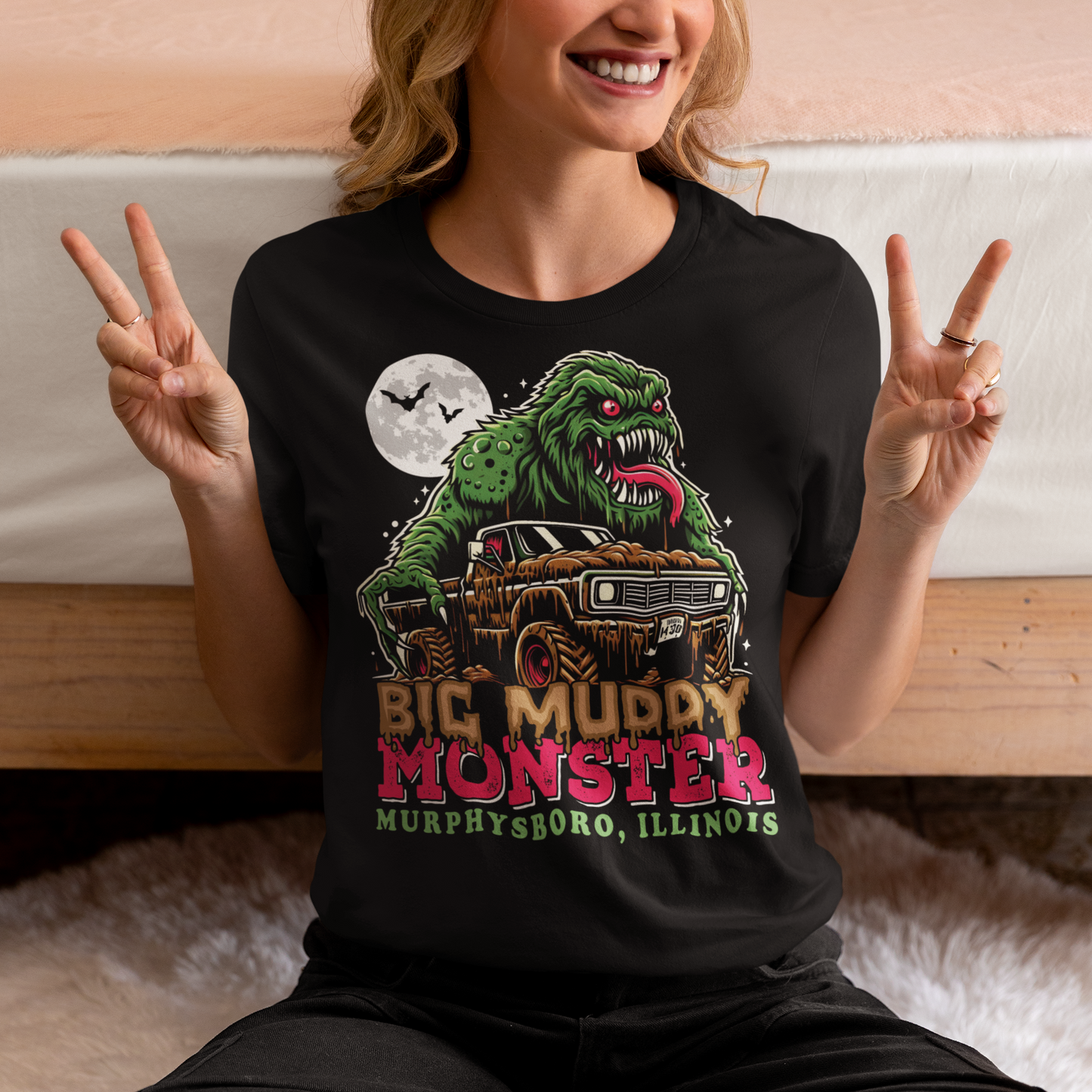 The t-shirt design features the Murphysboro Mud Monster, a legendary cryptid, attacking a rugged, lifted truck. The monster, depicted with a hulking, muddy, and shaggy appearance, looms large over the vehicle, gripping the sides with massive, clawed hands. Mud splatters dramatically across the truck's tires and fenders, capturing the wild essence of the creature. Below the scene, the bold text "BIG MUDDY MONSTER" is prominently displayed.
