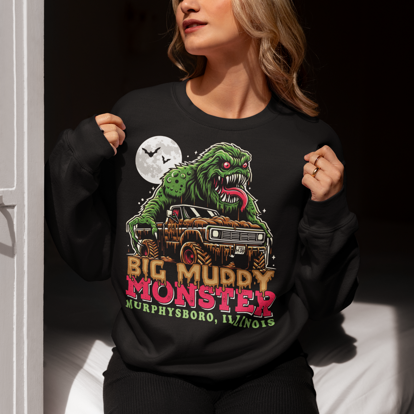 The sweatshirt design features the Murphysboro Mud Monster, a legendary cryptid, attacking a rugged, lifted truck. The monster, depicted with a hulking, muddy, and shaggy appearance, looms large over the vehicle, gripping the sides with massive, clawed hands. Mud splatters dramatically across the truck's tires and fenders, capturing the wild essence of the creature. Below the scene, the bold text "BIG MUDDY MONSTER" is prominently displayed.