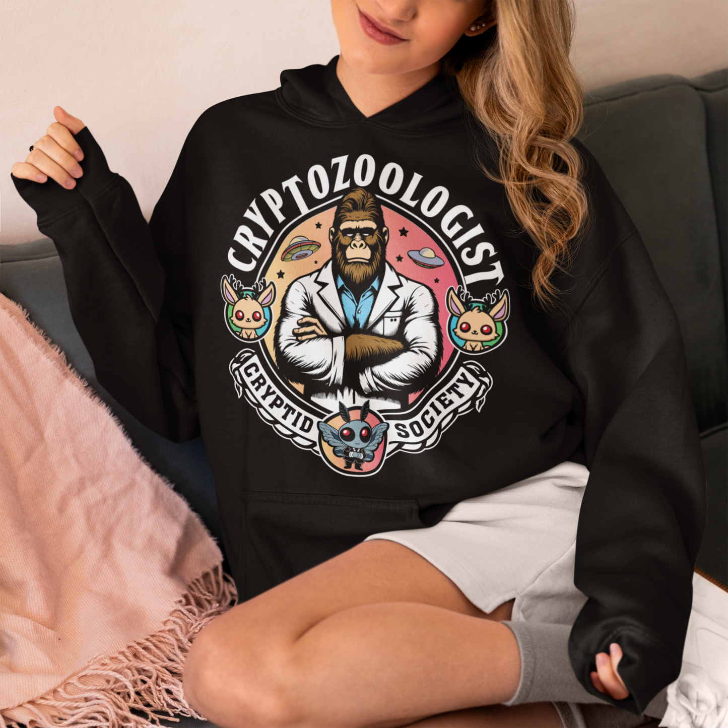 Black hoodie of a Bigfoot scientist with UFOs in the background. Mothman underneath and text above CRYPTOZOOLOGIST.