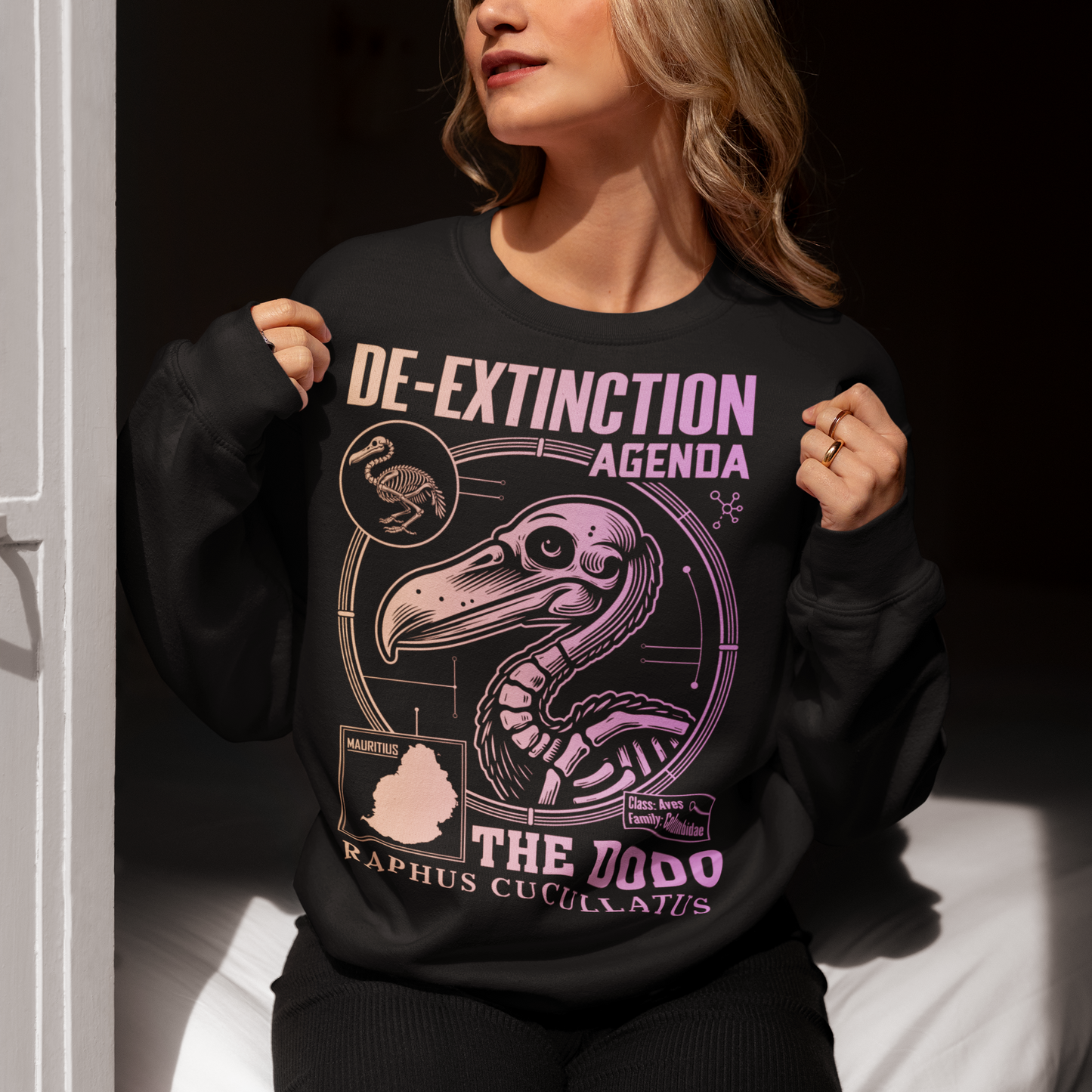 A graphic sweatshirt featuring a stylized illustration of a dodo bird, standing upright with a proud, almost defiant expression. The dodo is depicted in vibrant colors, blending realism with a slightly cartoonish flair. Above the bird, bold, capitalized text reads "DE-EXTINCTION AGENDA" in a futuristic font, suggesting a science fiction or activist tone. The design evokes themes of genetic engineering, conservation, and the revival of extinct species. The shirt is a statement piece with humor.