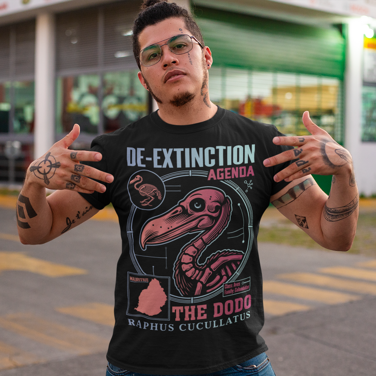A graphic t-shirt featuring a stylized illustration of a dodo bird, standing upright with a proud, almost defiant expression. The dodo is depicted in vibrant colors, blending realism with a slightly cartoonish flair. Above the bird, bold, capitalized text reads "DE-EXTINCTION AGENDA" in a futuristic font, suggesting a science fiction or activist tone. The design evokes themes of genetic engineering, conservation, and the revival of extinct species. The shirt is a statement piece with humor.