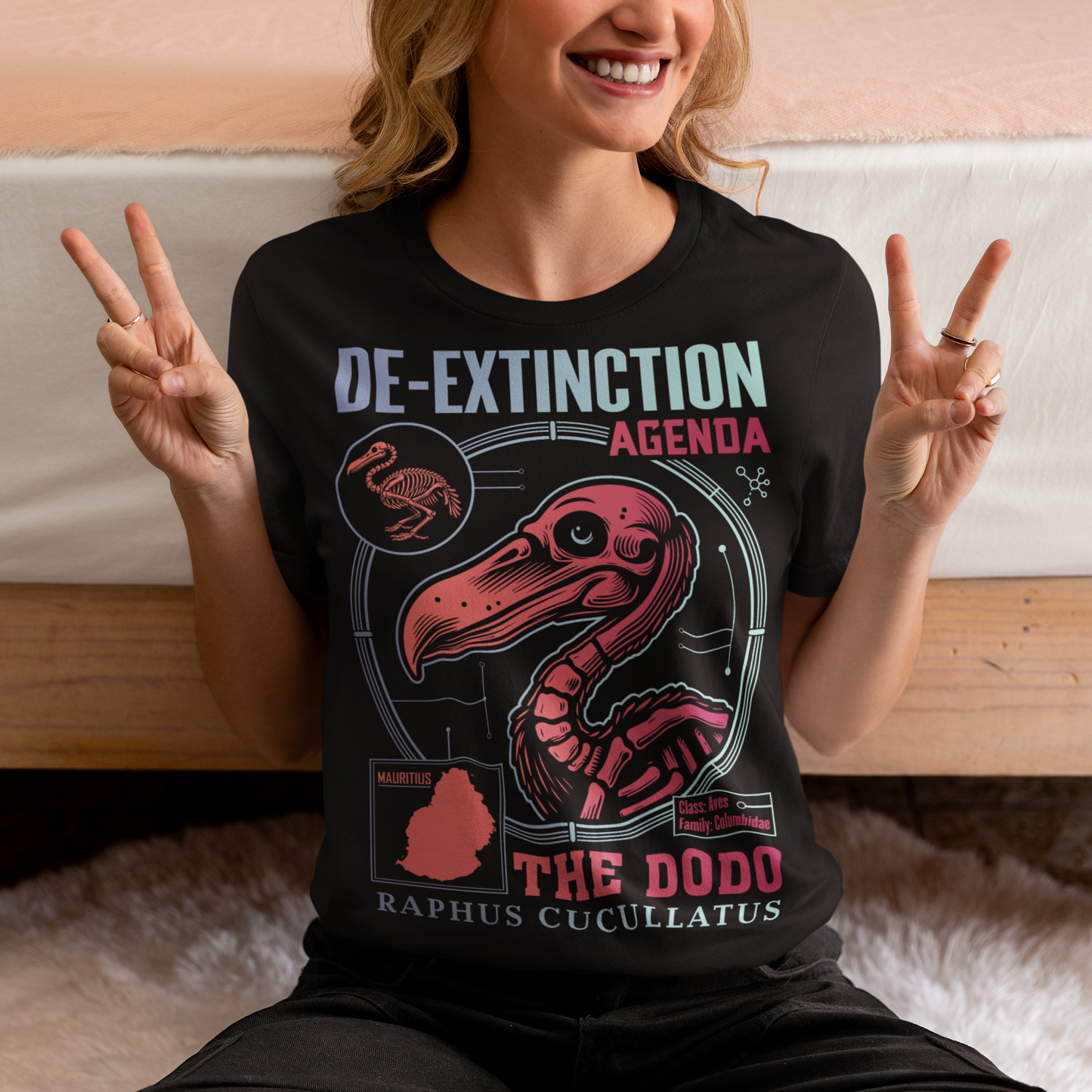 A graphic t-shirt featuring a stylized illustration of a dodo bird, standing upright with a proud, almost defiant expression. The dodo is depicted in vibrant colors, blending realism with a slightly cartoonish flair. Above the bird, bold, capitalized text reads "DE-EXTINCTION AGENDA" in a futuristic font, suggesting a science fiction or activist tone. The design evokes themes of genetic engineering, conservation, and the revival of extinct species. The shirt is a statement piece with humor.