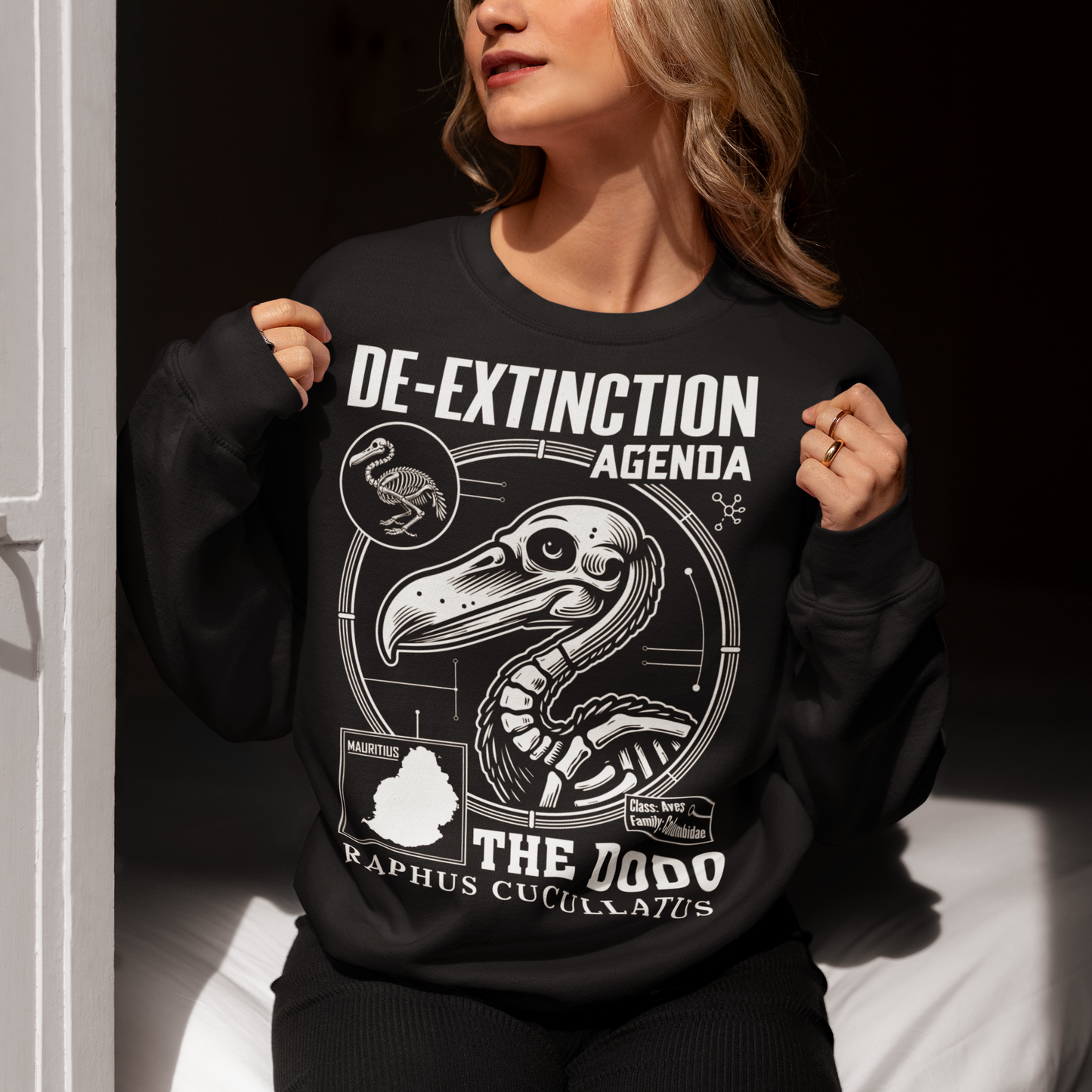 A graphic sweatshirt featuring a stylized illustration of a dodo bird, standing upright with a proud, almost defiant expression. The dodo is depicted in vibrant colors, blending realism with a slightly cartoonish flair. Above the bird, bold, capitalized text reads "DE-EXTINCTION AGENDA" in a futuristic font, suggesting a science fiction or activist tone. The design evokes themes of genetic engineering, conservation, and the revival of extinct species. The shirt is a statement piece with humor.
