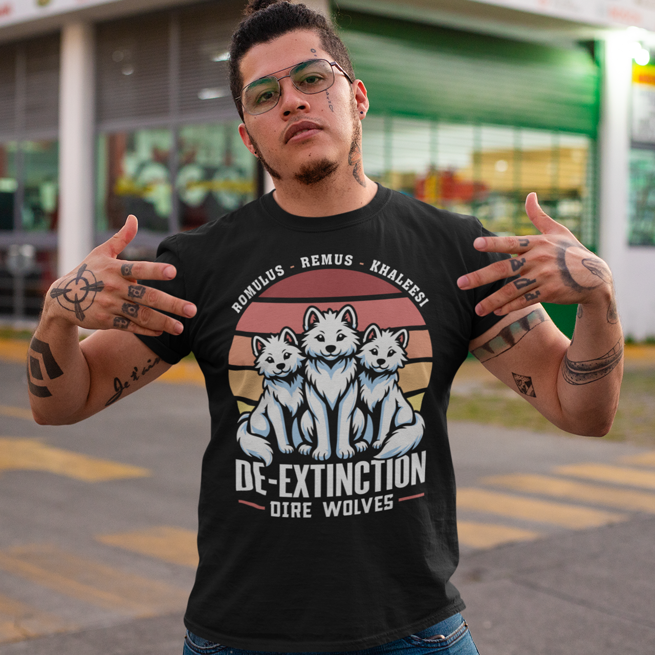 A graphic t-shirt design featuring three majestic Dire Wolves sitting side by side, facing forward with alert expressions. Behind them is a large, stylized retro sun composed of horizontal stripes in warm tones of orange, red, and yellow, evoking a vintage 80s aesthetic. Beneath the wolves, bold text reads “DE-EXTINCTION,” suggesting the return of these prehistoric predators. The design blends prehistoric imagery with nostalgic retro-futurism.
