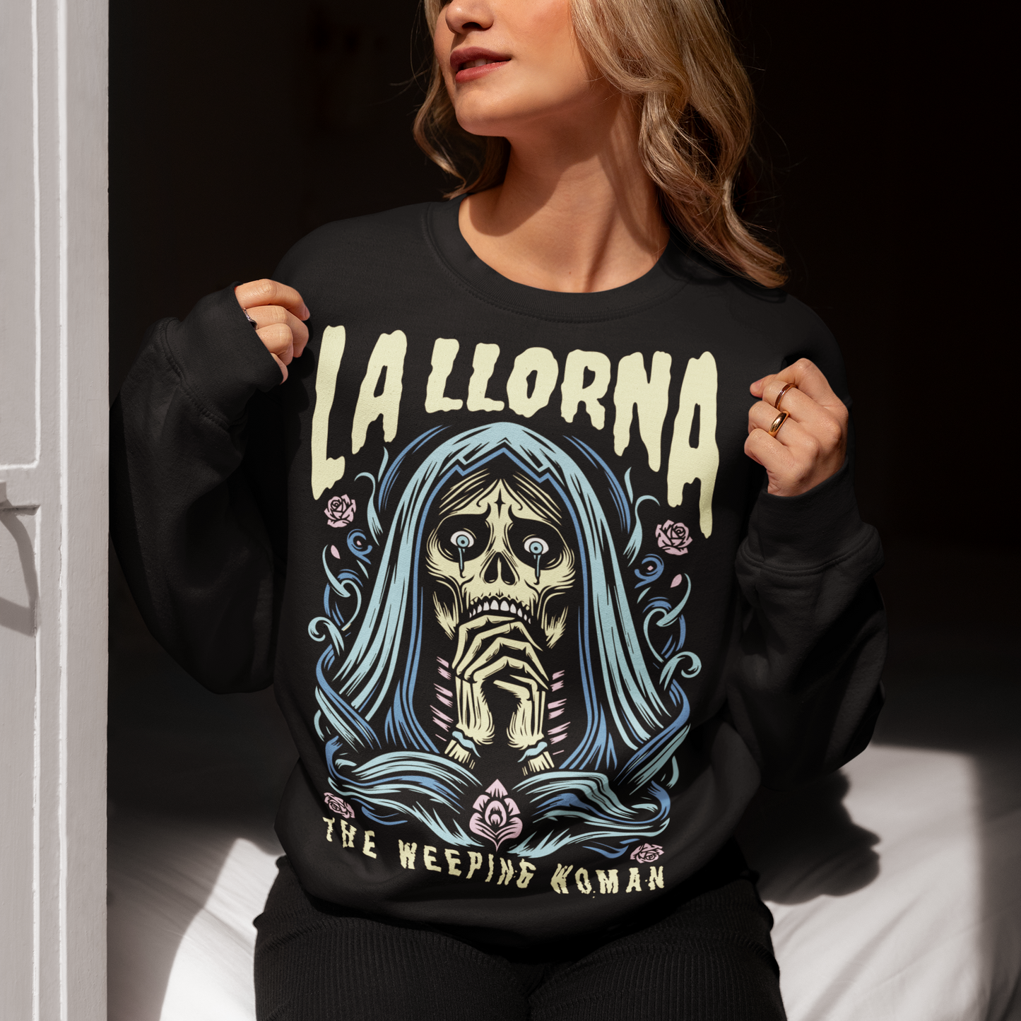 This striking La Llorona sweatshirt features a haunting depiction of the legendary Weeping Woman, her ghostly figure shrouded in mist, with flowing hair and a sorrowful expression as she searches for her lost children. The design captures the eerie, chilling essence of the famous Latin American folktale, blending dark, gothic tones with an ethereal glow. Above the image, bold lettering reads "LA LLORONA", while below, the translation "THE WEEPING WOMAN" adds to the eerie mystique.