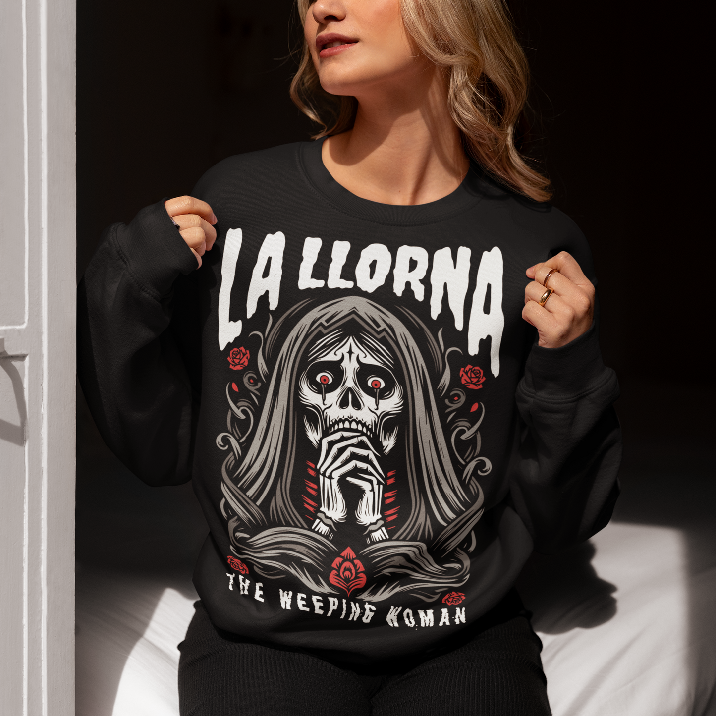 This striking La Llorona sweatshirt features a haunting depiction of the legendary Weeping Woman, her ghostly figure shrouded in mist, with flowing hair and a sorrowful expression as she searches for her lost children. The design captures the eerie, chilling essence of the famous Latin American folktale, blending dark, gothic tones with an ethereal glow. Above the image, bold lettering reads "LA LLORONA", while below, the translation "THE WEEPING WOMAN" adds to the eerie mystique.