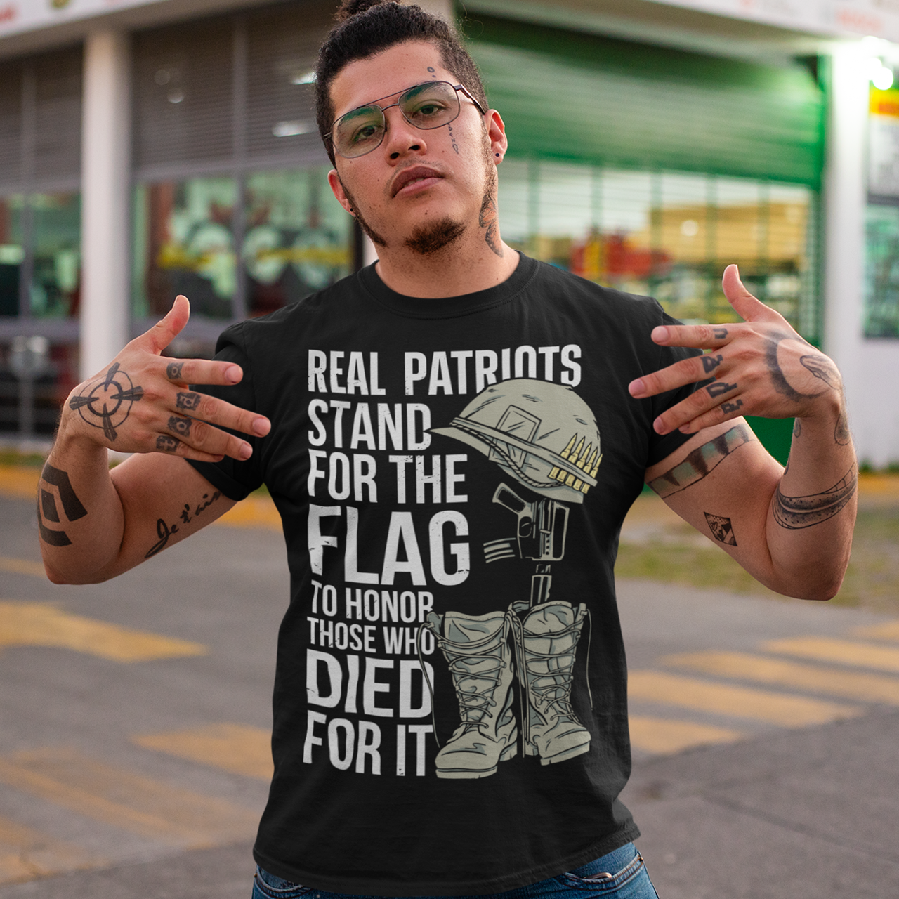 -shirt design featuring a graphic of a military helmet resting on a rifle, which is standing upright in a pair of combat boots—a battlefield cross symbolizing fallen soldiers. Above and below the image is bold text that reads: "REAL PATRIOTS STAND FOR THE FLAG TO HONOR THOSE WHO DIED FOR IT." The design uses patriotic imagery to emphasize respect and remembrance for military service members who have sacrificed their lives.