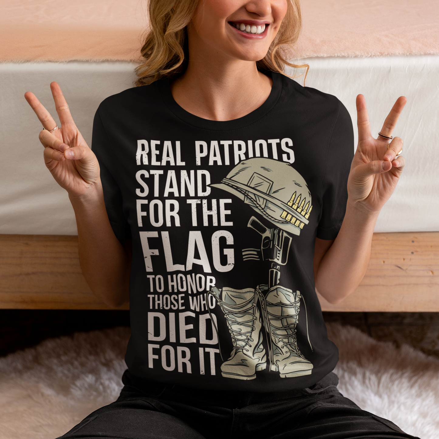 -shirt design featuring a graphic of a military helmet resting on a rifle, which is standing upright in a pair of combat boots—a battlefield cross symbolizing fallen soldiers. Above and below the image is bold text that reads: "REAL PATRIOTS STAND FOR THE FLAG TO HONOR THOSE WHO DIED FOR IT." The design uses patriotic imagery to emphasize respect and remembrance for military service members who have sacrificed their lives.