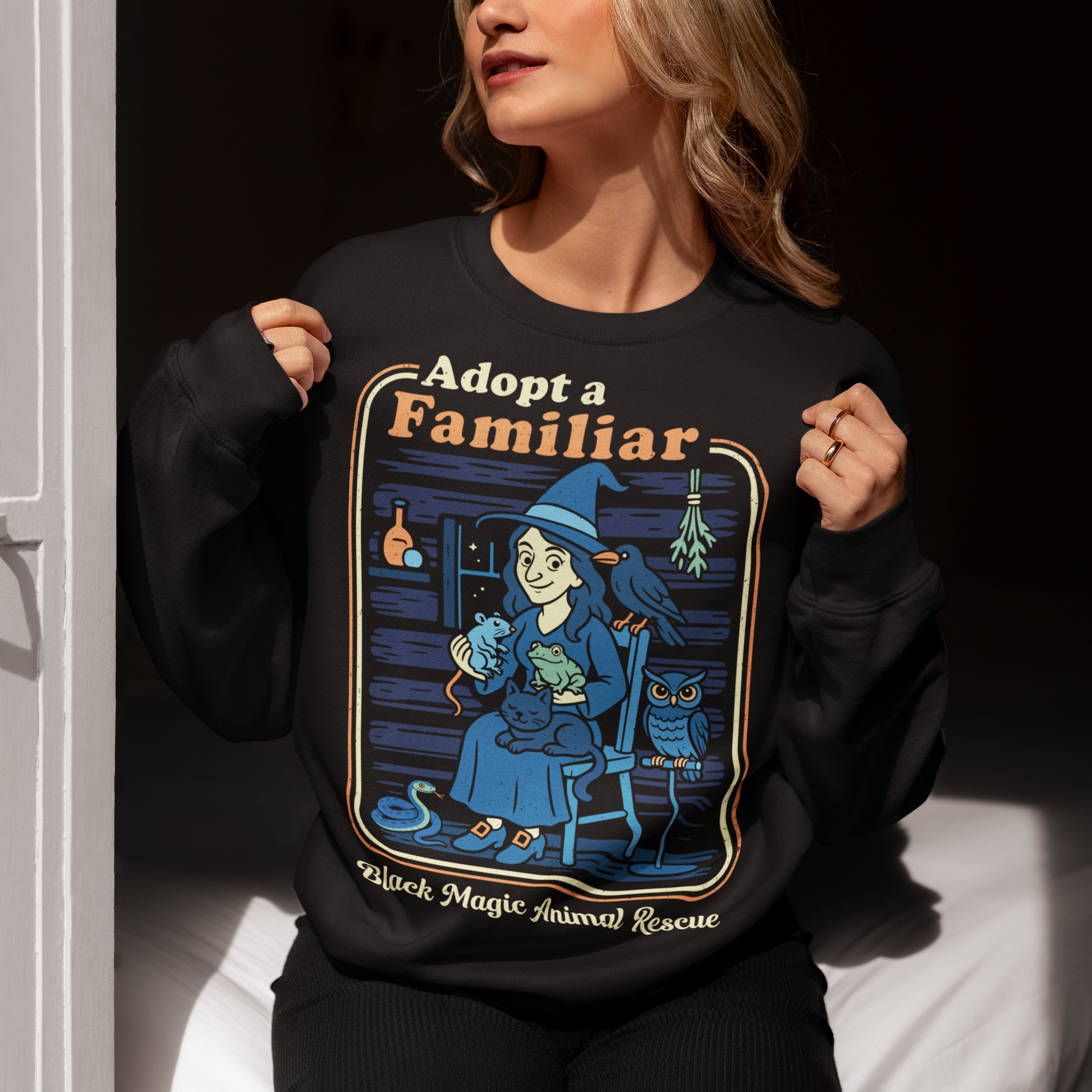 This cozy black sweatshirt features a charming illustration of a gentle witch surrounded by her rescued “familiars,” a cat curled in her lap, a curious frog, a loyal crow, a wise owl, and even a tiny snake slithering by. Framed like a vintage storybook scene, the design pairs warm, muted colors with playful linework and the heart-stealing phrase “Adopt a Familiar” above “Black Magic Animal Rescue. This shirt is perfect for animal lovers.