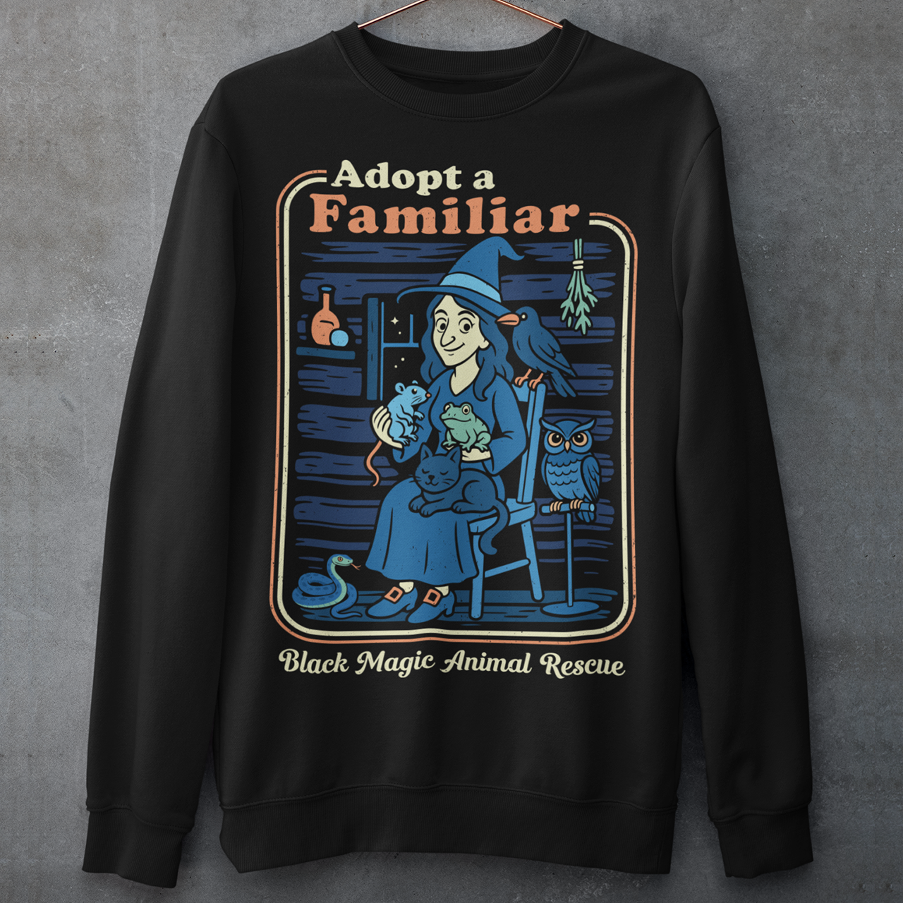 This cozy black sweatshirt features a charming illustration of a gentle witch surrounded by her rescued “familiars,” a cat curled in her lap, a curious frog, a loyal crow, a wise owl, and even a tiny snake slithering by. Framed like a vintage storybook scene, the design pairs warm, muted colors with playful linework and the heart-stealing phrase “Adopt a Familiar” above “Black Magic Animal Rescue. This shirt is perfect for animal lovers.
