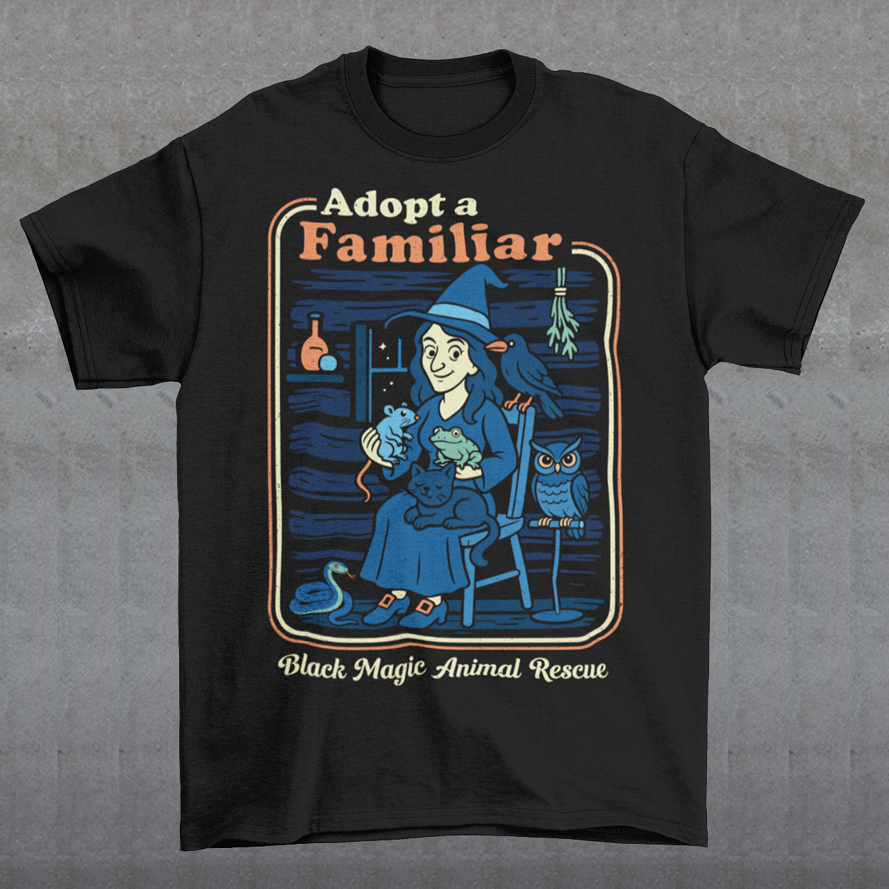 This cozy black t-shirt features a charming illustration of a gentle witch surrounded by her rescued “familiars,” a cat curled in her lap, a curious frog, a loyal crow, a wise owl, and even a tiny snake slithering by. Framed like a vintage storybook scene, the design pairs warm, muted colors with playful linework and the heart-stealing phrase “Adopt a Familiar” above “Black Magic Animal Rescue. This shirt is perfect for animal lovers.