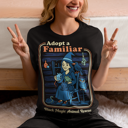 This cozy black t-shirt features a charming illustration of a gentle witch surrounded by her rescued “familiars,” a cat curled in her lap, a curious frog, a loyal crow, a wise owl, and even a tiny snake slithering by. Framed like a vintage storybook scene, the design pairs warm, muted colors with playful linework and the heart-stealing phrase “Adopt a Familiar” above “Black Magic Animal Rescue. This shirt is perfect for animal lovers.