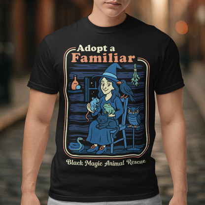 This cozy black t-shirt features a charming illustration of a gentle witch surrounded by her rescued “familiars,” a cat curled in her lap, a curious frog, a loyal crow, a wise owl, and even a tiny snake slithering by. Framed like a vintage storybook scene, the design pairs warm, muted colors with playful linework and the heart-stealing phrase “Adopt a Familiar” above “Black Magic Animal Rescue. This shirt is perfect for animal lovers.