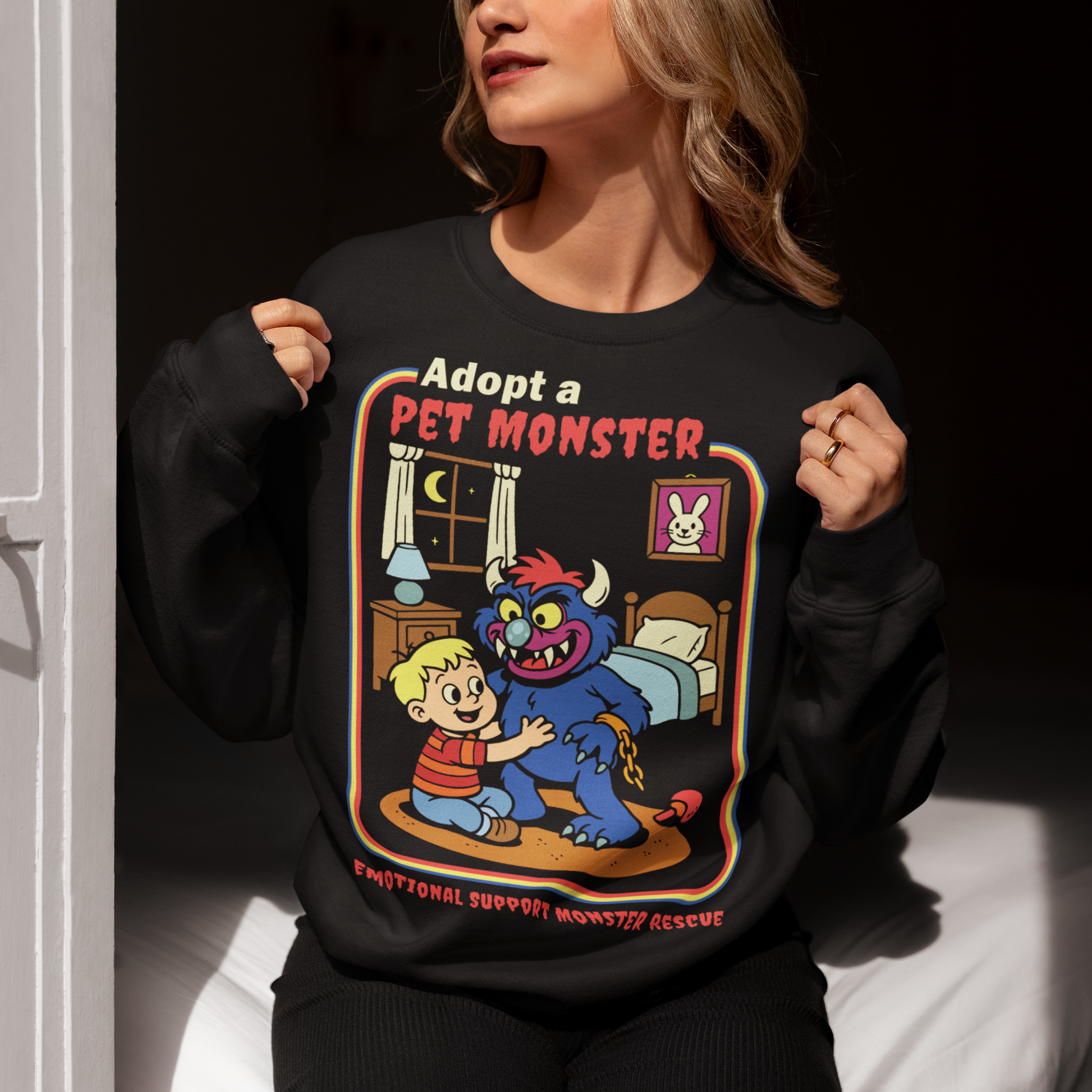 Soft, spooky, and suspiciously adorable, this sweatshirt looks like it came straight from a slightly haunted Saturday morning cartoon vault. The design features a wide eyed kid befriending a fuzzy little monster with tiny horns and big attitude, all set inside a cozy bedroom scene with retro storybook vibes. The bold “Adopt a Pet Monster” headline and “Emotional Support Monster Rescue” tagline sell the joke perfectly, blending cute chaos with feel good humor.