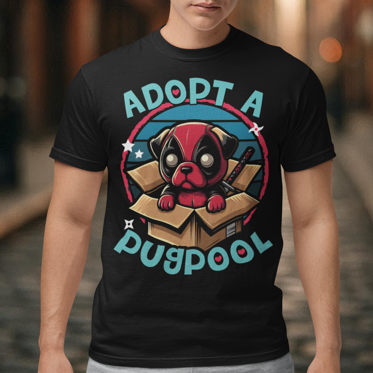 The T-shirt design features an adorable pug dressed as a mercenary, complete with a tiny red and black costume and a mask, sitting comfortably inside a slightly worn cardboard box. Around the design says "ADOPT A PUGPOOL" in a playful, hand-drawn font, giving it a quirky, homemade look.