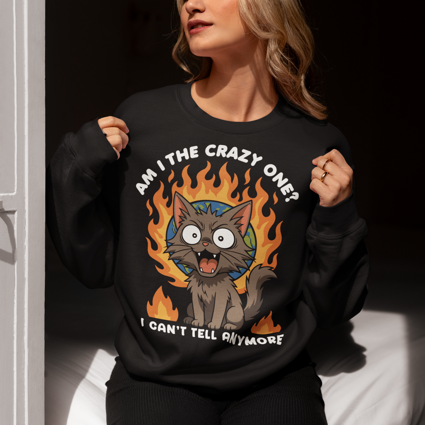 Black sweatshirt featuring a cartoon gray cat with wide eyes and sharp teeth, sitting in front of a flaming globe. The design includes the text “Am I the crazy one? I can’t tell anymore” in bold white letters. The illustration has a humorous, dark tone, combining chaotic energy with a cute yet slightly unhinged cat character.