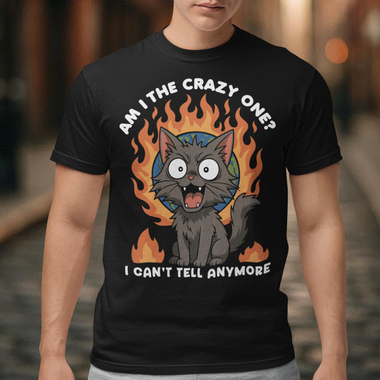 Black t-shirt featuring a cartoon gray cat with wide eyes and sharp teeth, sitting in front of a flaming globe. The design includes the text “Am I the crazy one? I can’t tell anymore” in bold white letters. The illustration has a humorous, dark tone, combining chaotic energy with a cute yet slightly unhinged cat character.