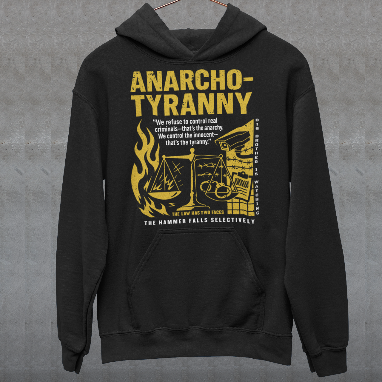 Black hoodie featuring a distressed yellow “ANARCHO-TYRANNY” graphic with scales of justice, flames, barbed wire, surveillance imagery, and prison-themed artwork. The design includes the phrases “THE LAW HAS TWO FACES” and “THE HAMMER FALLS SELECTIVELY,” creating a bold political statement focused on authority, government power, civil liberties, and anti-authoritarian themes.