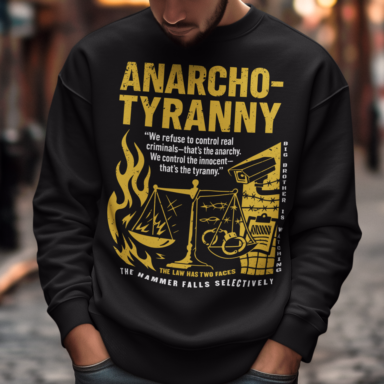 Black sweatshirt featuring a distressed yellow “ANARCHO-TYRANNY” graphic with scales of justice, flames, barbed wire, surveillance imagery, and prison-themed artwork. The design includes the phrases “THE LAW HAS TWO FACES” and “THE HAMMER FALLS SELECTIVELY,” creating a bold political statement focused on authority, government power, civil liberties, and anti-authoritarian themes.