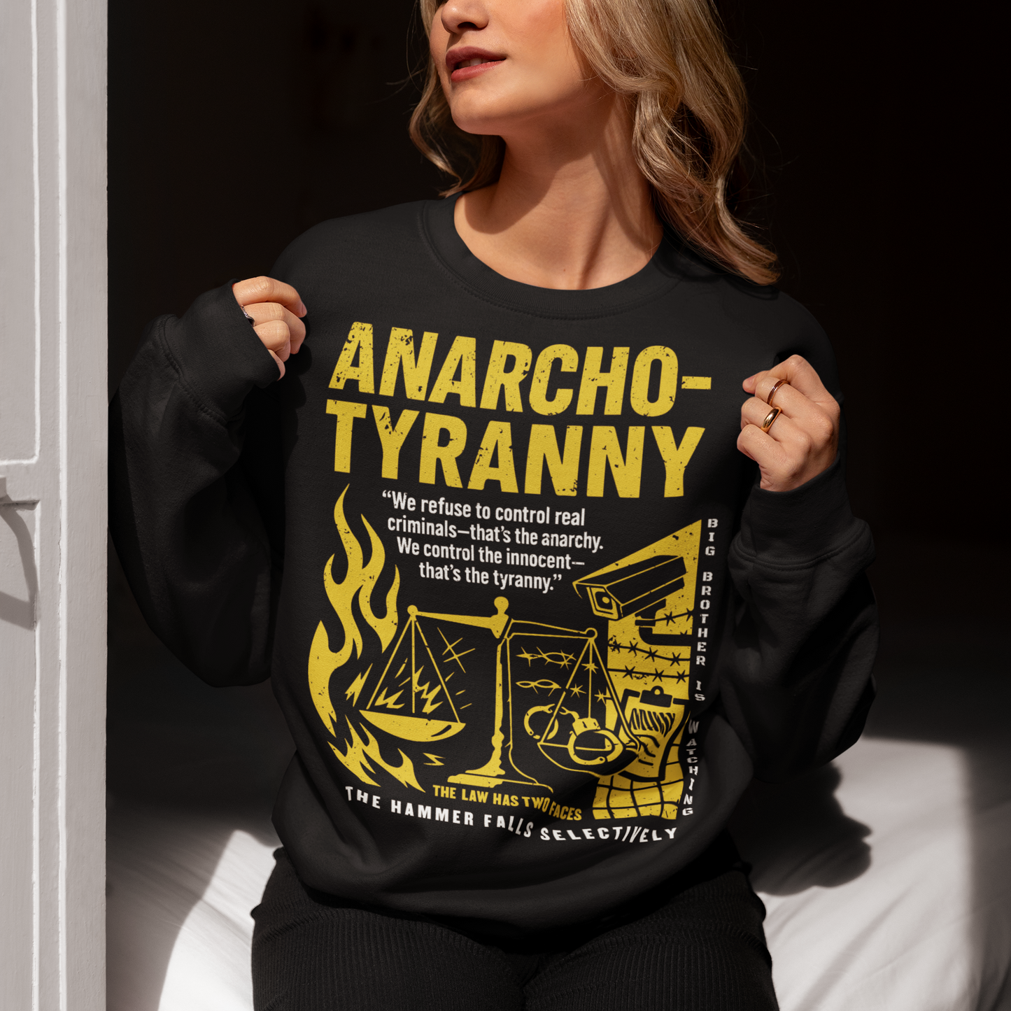 Black sweatshirt featuring a distressed yellow “ANARCHO-TYRANNY” graphic with scales of justice, flames, barbed wire, surveillance imagery, and prison-themed artwork. The design includes the phrases “THE LAW HAS TWO FACES” and “THE HAMMER FALLS SELECTIVELY,” creating a bold political statement focused on authority, government power, civil liberties, and anti-authoritarian themes.