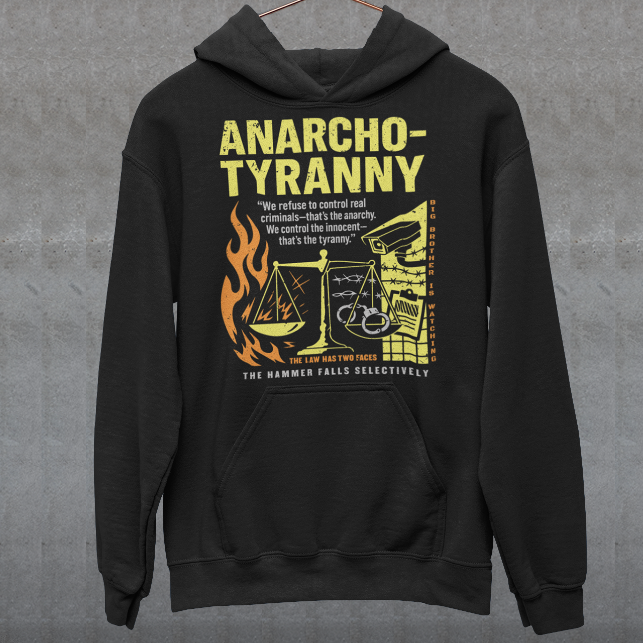 Black hoodie featuring a distressed yellow “ANARCHO-TYRANNY” graphic with scales of justice, flames, barbed wire, surveillance imagery, and prison-themed artwork. The design includes the phrases “THE LAW HAS TWO FACES” and “THE HAMMER FALLS SELECTIVELY,” creating a bold political statement focused on authority, government power, civil liberties, and anti-authoritarian themes.
