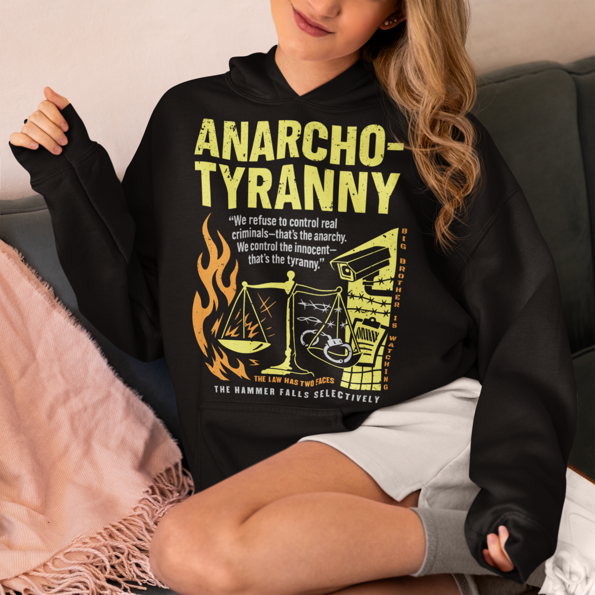 Black hoodie featuring a distressed yellow “ANARCHO-TYRANNY” graphic with scales of justice, flames, barbed wire, surveillance imagery, and prison-themed artwork. The design includes the phrases “THE LAW HAS TWO FACES” and “THE HAMMER FALLS SELECTIVELY,” creating a bold political statement focused on authority, government power, civil liberties, and anti-authoritarian themes.