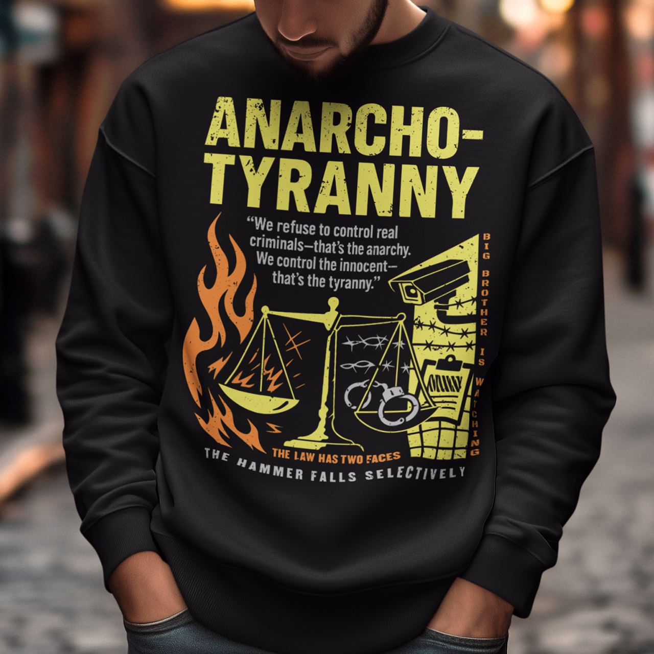 Black sweatshirt featuring a distressed yellow “ANARCHO-TYRANNY” graphic with scales of justice, flames, barbed wire, surveillance imagery, and prison-themed artwork. The design includes the phrases “THE LAW HAS TWO FACES” and “THE HAMMER FALLS SELECTIVELY,” creating a bold political statement focused on authority, government power, civil liberties, and anti-authoritarian themes.