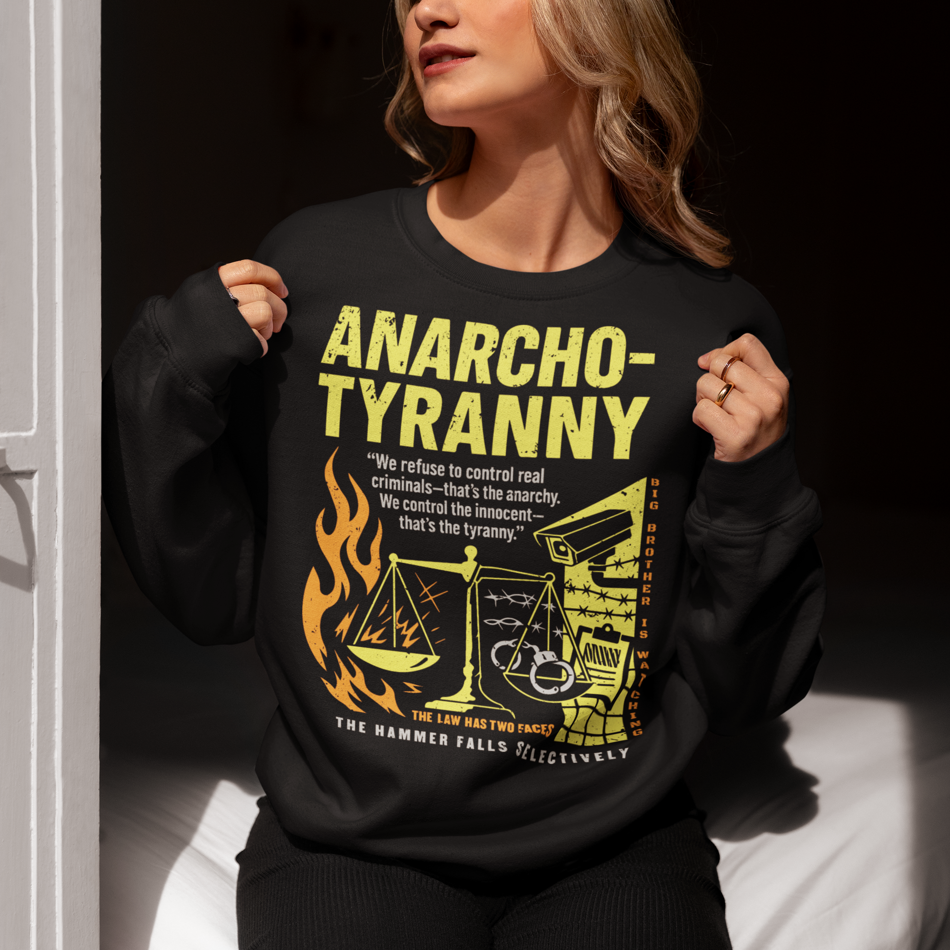 Black sweatshirt featuring a distressed yellow “ANARCHO-TYRANNY” graphic with scales of justice, flames, barbed wire, surveillance imagery, and prison-themed artwork. The design includes the phrases “THE LAW HAS TWO FACES” and “THE HAMMER FALLS SELECTIVELY,” creating a bold political statement focused on authority, government power, civil liberties, and anti-authoritarian themes.