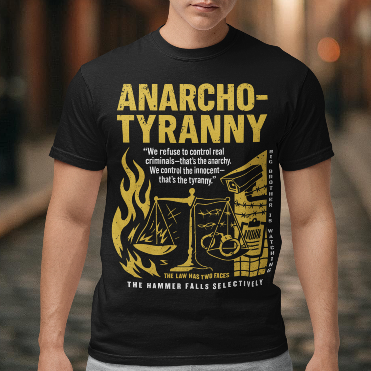 Black t-shirt featuring a distressed yellow “ANARCHO-TYRANNY” graphic with scales of justice, flames, barbed wire, surveillance imagery, and prison-themed artwork. The design includes the phrases “THE LAW HAS TWO FACES” and “THE HAMMER FALLS SELECTIVELY,” creating a bold political statement focused on authority, government power, civil liberties, and anti-authoritarian themes.