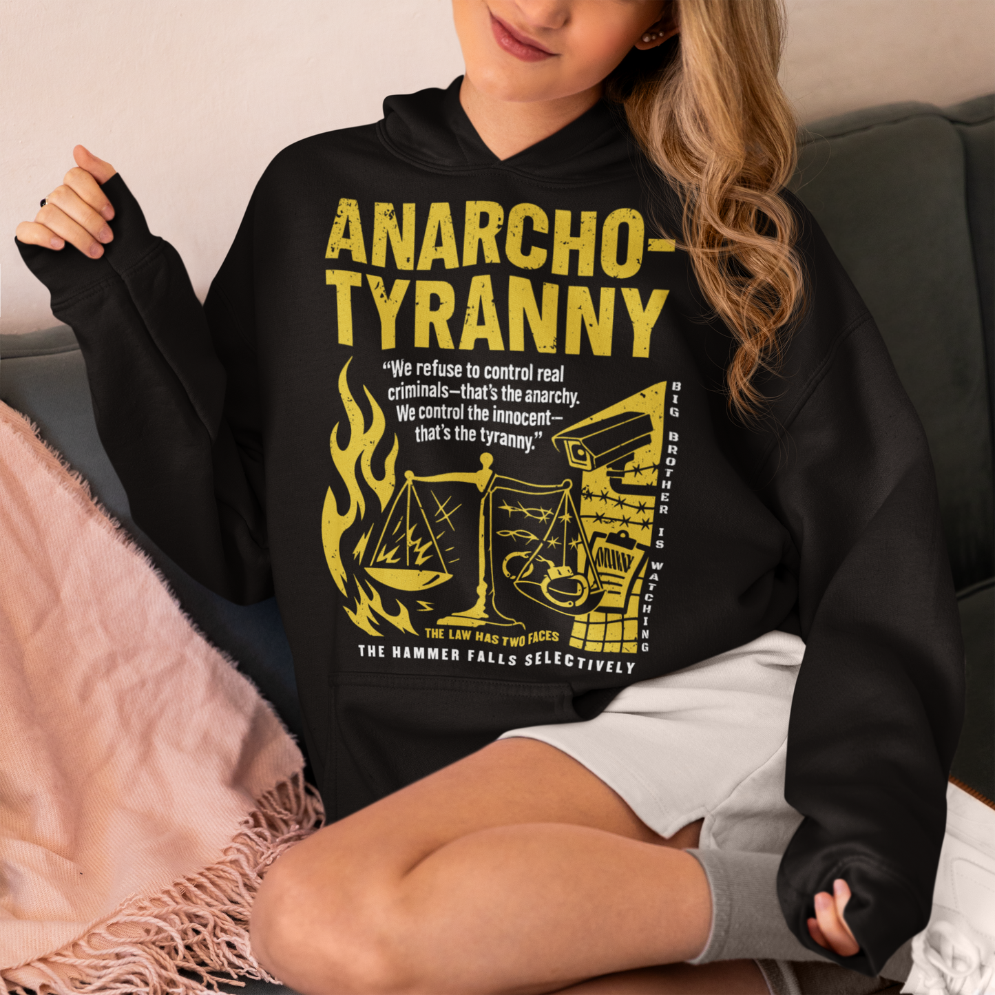 Black hoodie featuring a distressed yellow “ANARCHO-TYRANNY” graphic with scales of justice, flames, barbed wire, surveillance imagery, and prison-themed artwork. The design includes the phrases “THE LAW HAS TWO FACES” and “THE HAMMER FALLS SELECTIVELY,” creating a bold political statement focused on authority, government power, civil liberties, and anti-authoritarian themes.