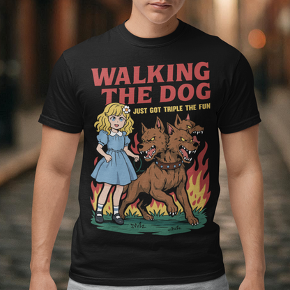 Black t-shirt featuring a colorful cartoon design of a young girl in a blue dress casually walking a three-headed Cerberus dog on a leash. The fierce hellhound snarls with sharp teeth while flames rise behind it, creating a striking contrast between cute and chaotic. Bold red text reads “Walking the Dog” with smaller text below saying “Just Got Triple the Fun,” adding humor to the fantasy-inspired scene.