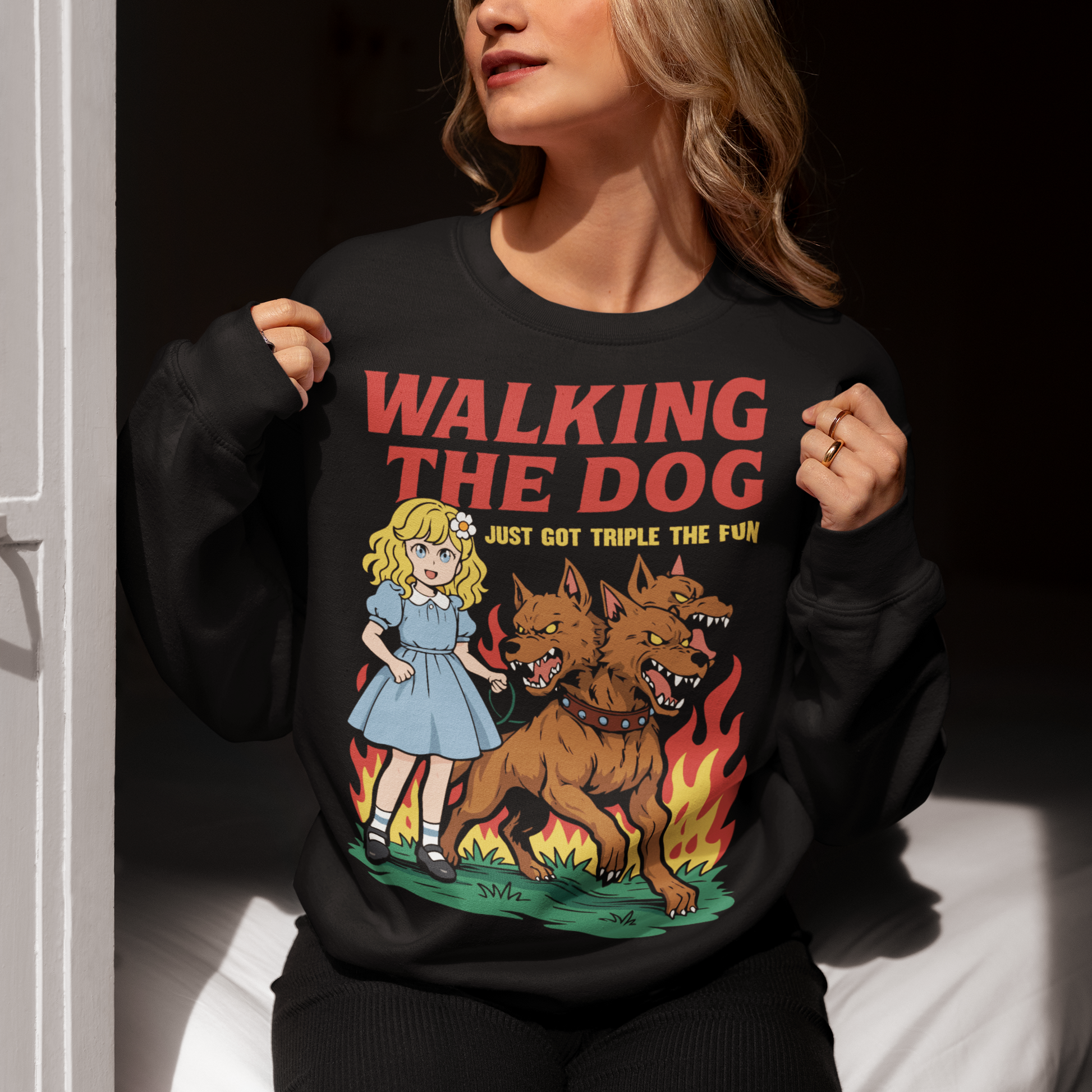 Black Sweatshirt featuring a colorful cartoon design of a young girl in a blue dress casually walking a three-headed Cerberus dog on a leash. The fierce hellhound snarls with sharp teeth while flames rise behind it, creating a striking contrast between cute and chaotic. Bold red text reads “Walking the Dog” with smaller text below saying “Just Got Triple the Fun,” adding humor to the fantasy-inspired scene.