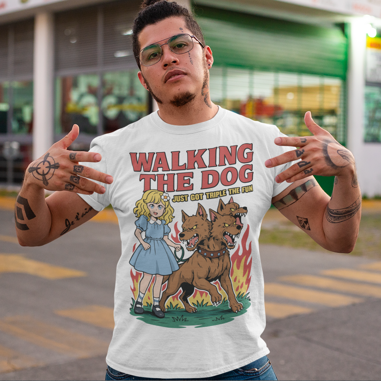 White t-shirt featuring a colorful cartoon design of a young girl in a blue dress casually walking a three-headed Cerberus dog on a leash. The fierce hellhound snarls with sharp teeth while flames rise behind it, creating a striking contrast between cute and chaotic. Bold red text reads “Walking the Dog” with smaller text below saying “Just Got Triple the Fun,” adding humor to the fantasy-inspired scene.