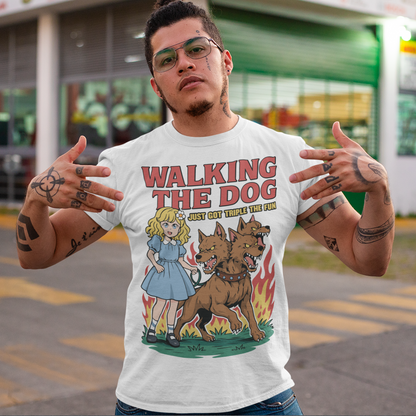 White t-shirt featuring a colorful cartoon design of a young girl in a blue dress casually walking a three-headed Cerberus dog on a leash. The fierce hellhound snarls with sharp teeth while flames rise behind it, creating a striking contrast between cute and chaotic. Bold red text reads “Walking the Dog” with smaller text below saying “Just Got Triple the Fun,” adding humor to the fantasy-inspired scene.