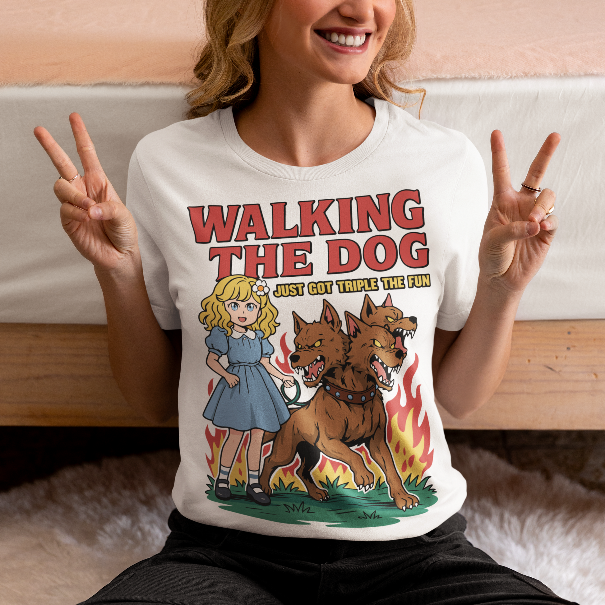 White t-shirt featuring a colorful cartoon design of a young girl in a blue dress casually walking a three-headed Cerberus dog on a leash. The fierce hellhound snarls with sharp teeth while flames rise behind it, creating a striking contrast between cute and chaotic. Bold red text reads “Walking the Dog” with smaller text below saying “Just Got Triple the Fun,” adding humor to the fantasy-inspired scene.