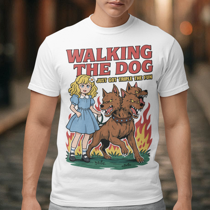 White t-shirt featuring a colorful cartoon design of a young girl in a blue dress casually walking a three-headed Cerberus dog on a leash. The fierce hellhound snarls with sharp teeth while flames rise behind it, creating a striking contrast between cute and chaotic. Bold red text reads “Walking the Dog” with smaller text below saying “Just Got Triple the Fun,” adding humor to the fantasy-inspired scene.
