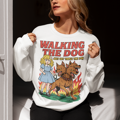 White Sweatshirt featuring a colorful cartoon design of a young girl in a blue dress casually walking a three-headed Cerberus dog on a leash. The fierce hellhound snarls with sharp teeth while flames rise behind it, creating a striking contrast between cute and chaotic. Bold red text reads “Walking the Dog” with smaller text below saying “Just Got Triple the Fun,” adding humor to the fantasy-inspired scene.