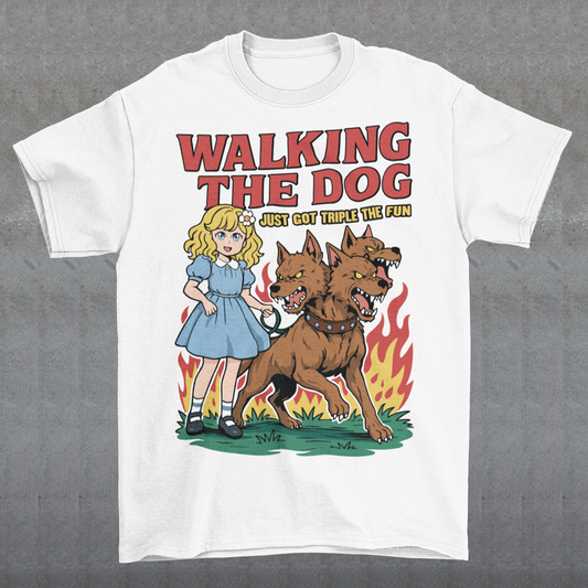 White t-shirt featuring a colorful cartoon design of a young girl in a blue dress casually walking a three-headed Cerberus dog on a leash. The fierce hellhound snarls with sharp teeth while flames rise behind it, creating a striking contrast between cute and chaotic. Bold red text reads “Walking the Dog” with smaller text below saying “Just Got Triple the Fun,” adding humor to the fantasy-inspired scene.