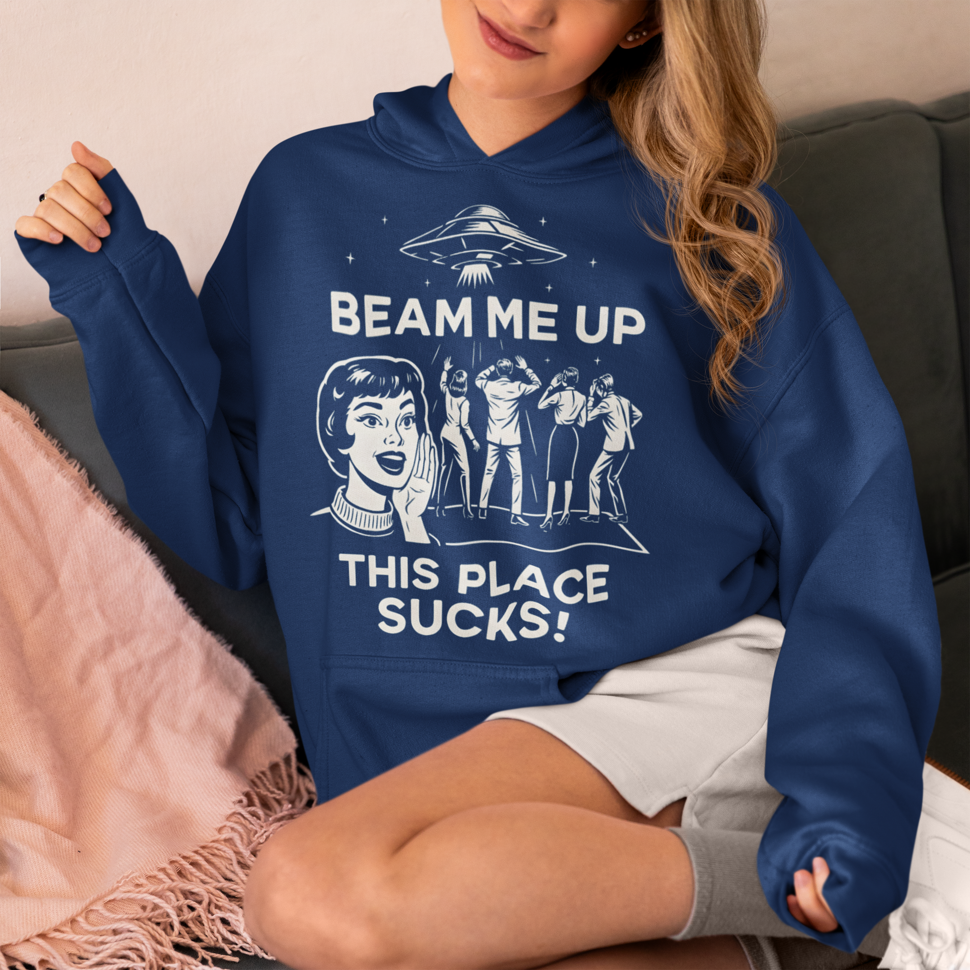 This snark-powered retro UFO hoodie brings the perfect escape fantasy to life. A vintage-style flying saucer hovers above a crowd while a wide-eyed woman rallies for interstellar rescue, paired with the bold line “Beam Me Up, This Place Sucks!” The design has a classic pulp-sci-fi look that feels like it tumbled out of an old comic rack. Ideal for anyone who survives their days with eye rolls, cosmic daydreams, and a desire to be anywhere else in the galaxy.
