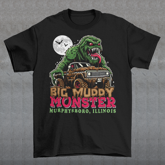 The t-shirt design features the Murphysboro Mud Monster, a legendary cryptid, attacking a rugged, lifted truck. The monster, depicted with a hulking, muddy, and shaggy appearance, looms large over the vehicle, gripping the sides with massive, clawed hands. Mud splatters dramatically across the truck's tires and fenders, capturing the wild essence of the creature. Below the scene, the bold text "BIG MUDDY MONSTER" is prominently displayed.