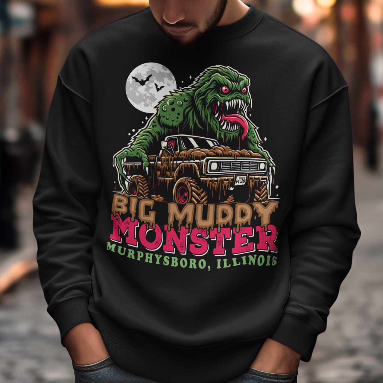 The sweatshirt design features the Murphysboro Mud Monster, a legendary cryptid, attacking a rugged, lifted truck. The monster, depicted with a hulking, muddy, and shaggy appearance, looms large over the vehicle, gripping the sides with massive, clawed hands. Mud splatters dramatically across the truck's tires and fenders, capturing the wild essence of the creature. Below the scene, the bold text "BIG MUDDY MONSTER" is prominently displayed.