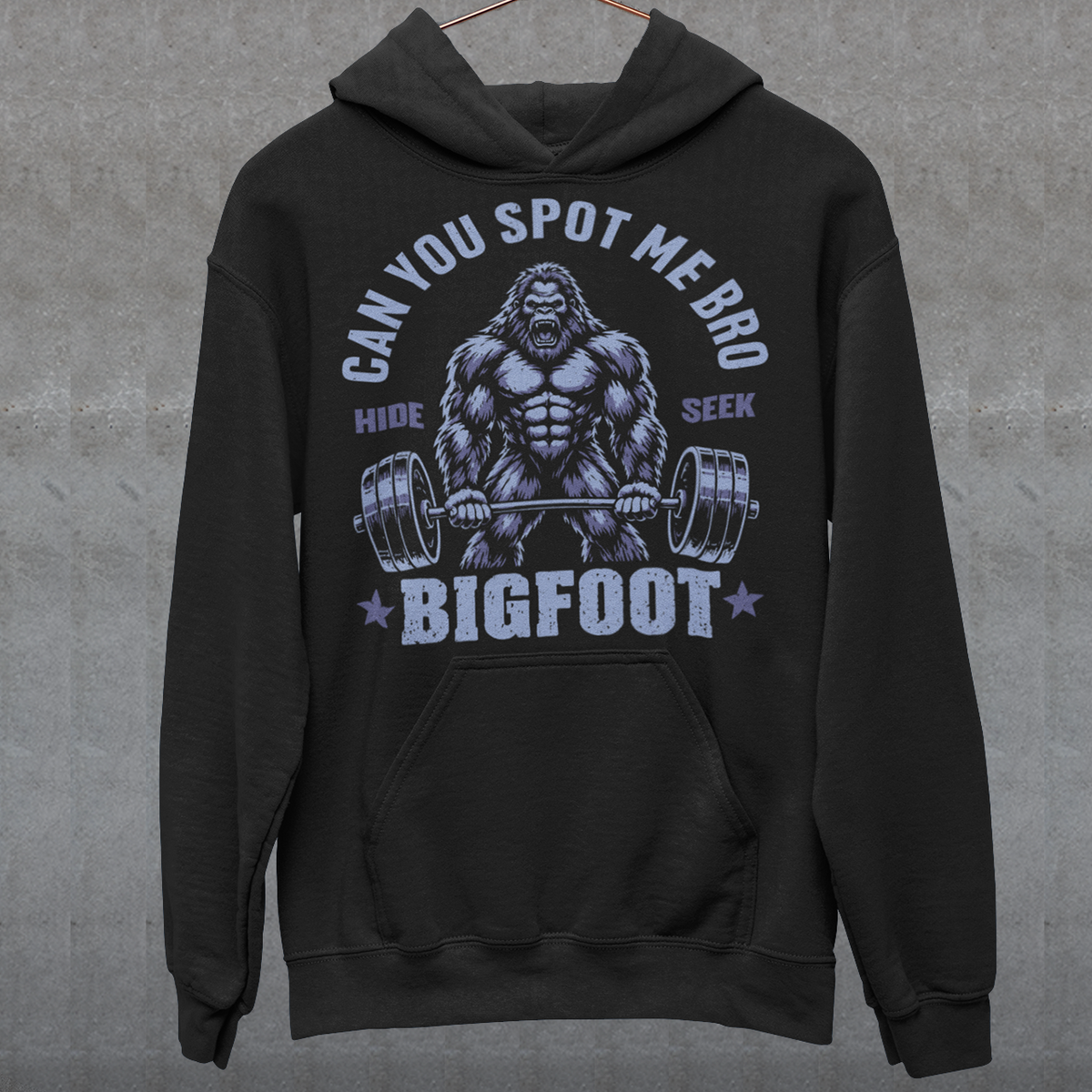 Black Hoodie featuring a muscular cartoon Bigfoot lifting a heavily loaded barbell. The graphic has bold white vintage-style lettering reading “CAN YOU SPOT ME BRO” above the Sasquatch, with “HIDE” and “SEEK” on either side and “BIGFOOT” beneath the weightlifter, flanked by stars. The design combines Bigfoot humor, gym culture, weightlifting, and cryptid themes.