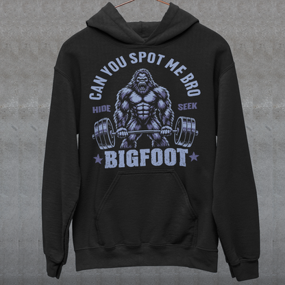Black Hoodie featuring a muscular cartoon Bigfoot lifting a heavily loaded barbell. The graphic has bold white vintage-style lettering reading “CAN YOU SPOT ME BRO” above the Sasquatch, with “HIDE” and “SEEK” on either side and “BIGFOOT” beneath the weightlifter, flanked by stars. The design combines Bigfoot humor, gym culture, weightlifting, and cryptid themes.
