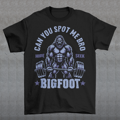 Black T-shirt featuring a muscular cartoon Bigfoot lifting a heavily loaded barbell. The graphic has bold white vintage-style lettering reading “CAN YOU SPOT ME BRO” above the Sasquatch, with “HIDE” and “SEEK” on either side and “BIGFOOT” beneath the weightlifter, flanked by stars. The design combines Bigfoot humor, gym culture, weightlifting, and cryptid themes.