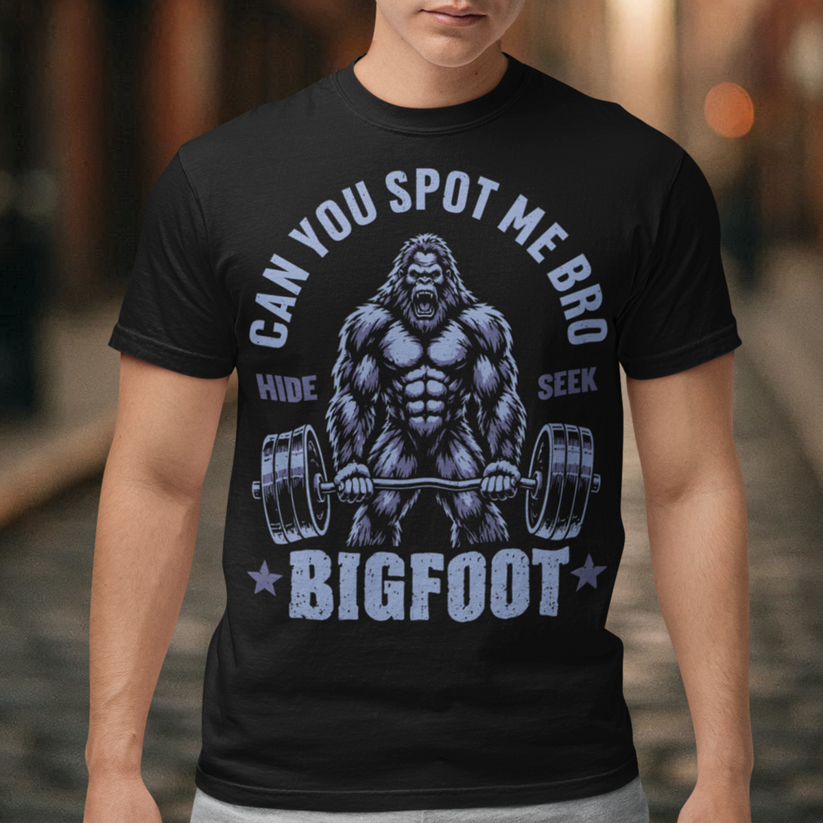 Black T-shirt featuring a muscular cartoon Bigfoot lifting a heavily loaded barbell. The graphic has bold white vintage-style lettering reading “CAN YOU SPOT ME BRO” above the Sasquatch, with “HIDE” and “SEEK” on either side and “BIGFOOT” beneath the weightlifter, flanked by stars. The design combines Bigfoot humor, gym culture, weightlifting, and cryptid themes.