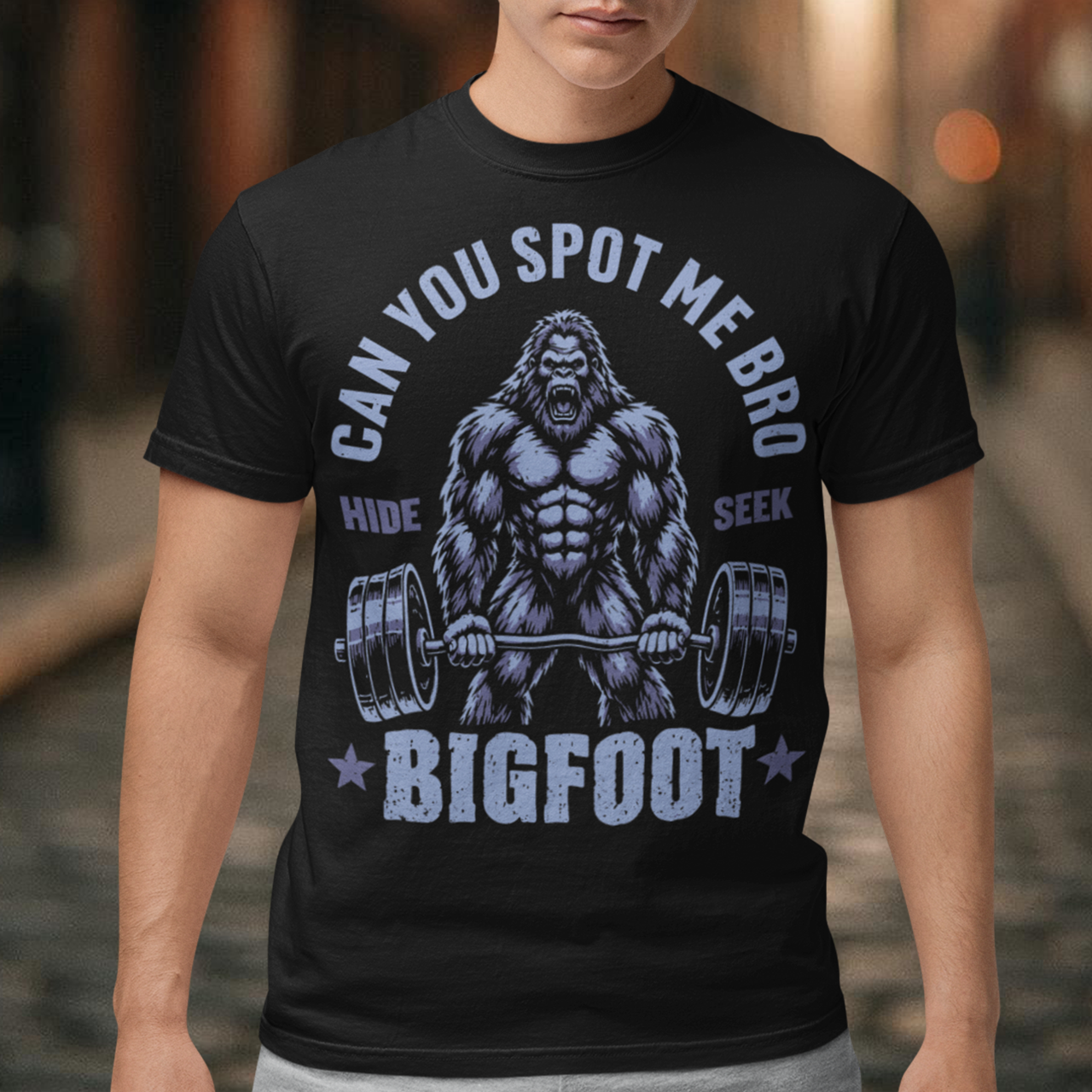 Black T-shirt featuring a muscular cartoon Bigfoot lifting a heavily loaded barbell. The graphic has bold white vintage-style lettering reading “CAN YOU SPOT ME BRO” above the Sasquatch, with “HIDE” and “SEEK” on either side and “BIGFOOT” beneath the weightlifter, flanked by stars. The design combines Bigfoot humor, gym culture, weightlifting, and cryptid themes.