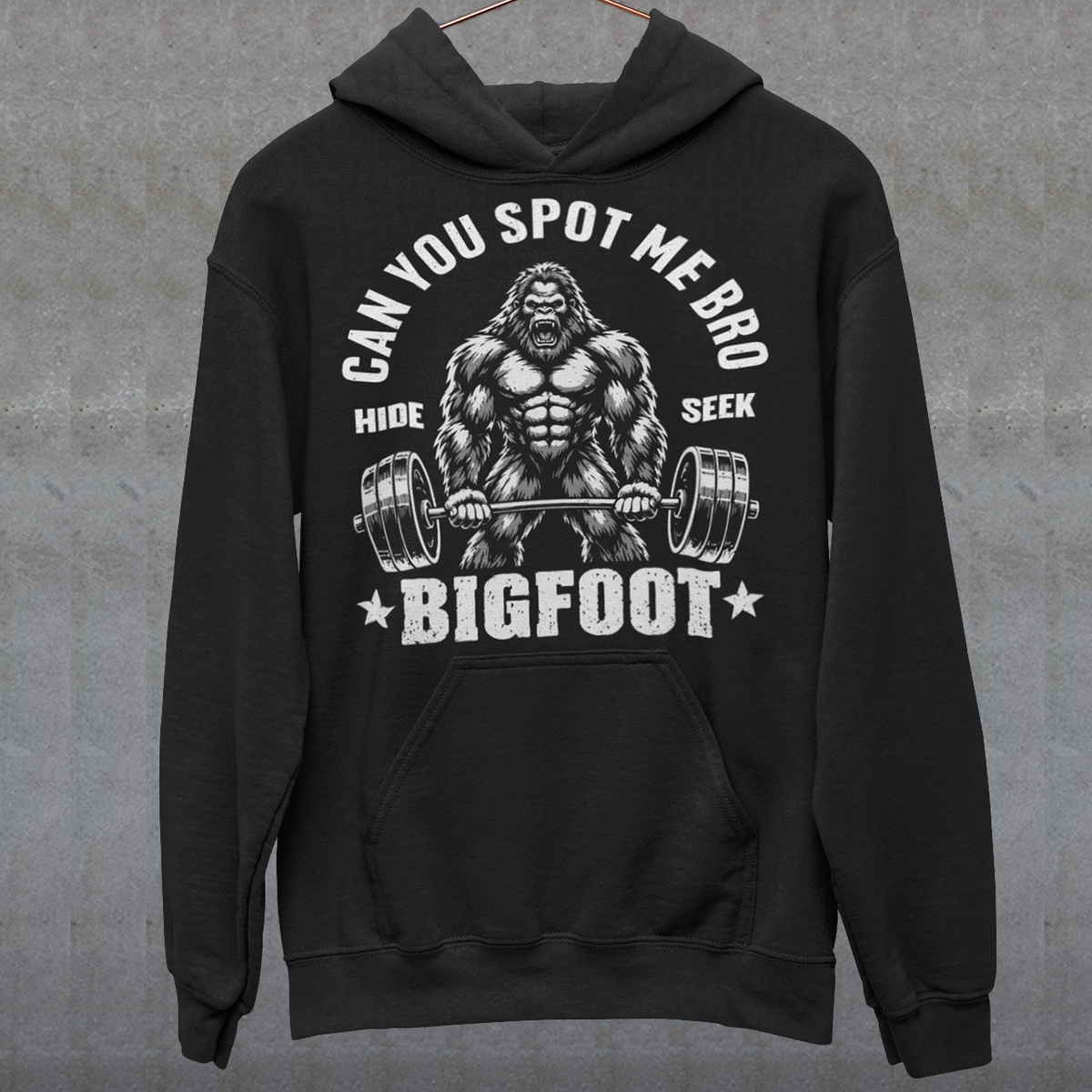 Black Hoodie featuring a muscular cartoon Bigfoot lifting a heavily loaded barbell. The graphic has bold white vintage-style lettering reading “CAN YOU SPOT ME BRO” above the Sasquatch, with “HIDE” and “SEEK” on either side and “BIGFOOT” beneath the weightlifter, flanked by stars. The design combines Bigfoot humor, gym culture, weightlifting, and cryptid themes.