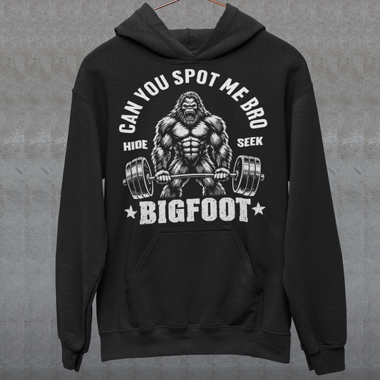 Black Hoodie featuring a muscular cartoon Bigfoot lifting a heavily loaded barbell. The graphic has bold white vintage-style lettering reading “CAN YOU SPOT ME BRO” above the Sasquatch, with “HIDE” and “SEEK” on either side and “BIGFOOT” beneath the weightlifter, flanked by stars. The design combines Bigfoot humor, gym culture, weightlifting, and cryptid themes.